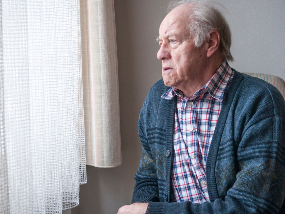 An older man in a blue cardigan and checked shirt sits at home, looking out through a net-curtained window.