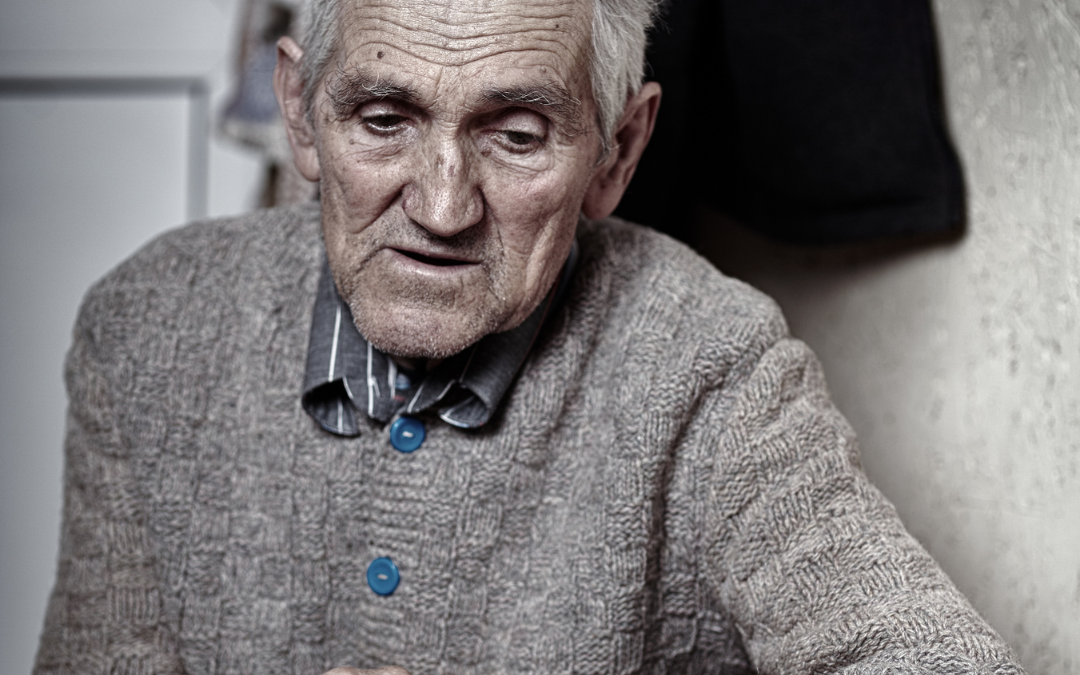 An older man in a knitted cardigan sits alone at a table, hands clasped, looking downwards