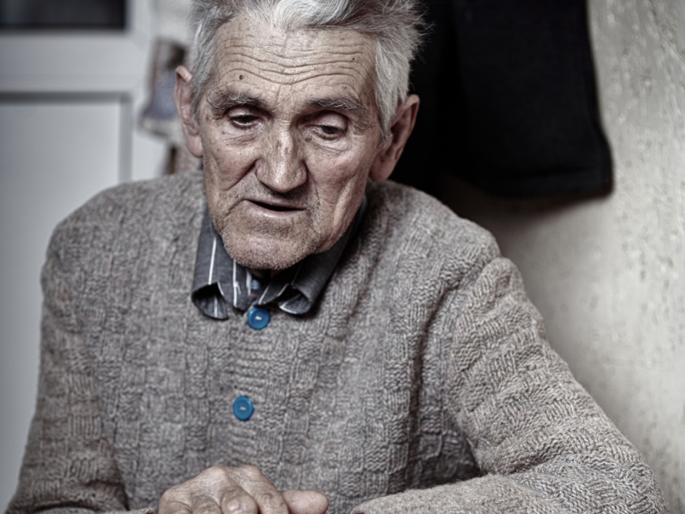 An older man in a knitted cardigan sits alone at a table, hands clasped, looking downwards