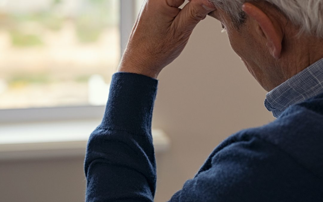 Older man in a navy jumper sits by a window with his hand to his forehead, looking downcast