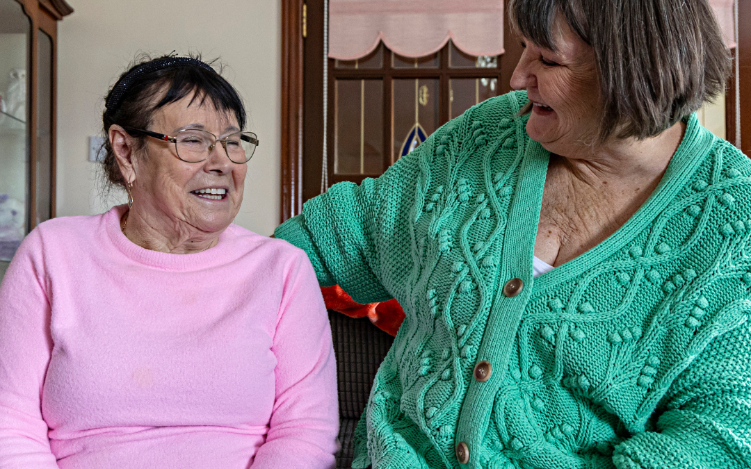 Two women laughing together on a sofa in a bright sitting room, one in pink, the other in a green cardigan
