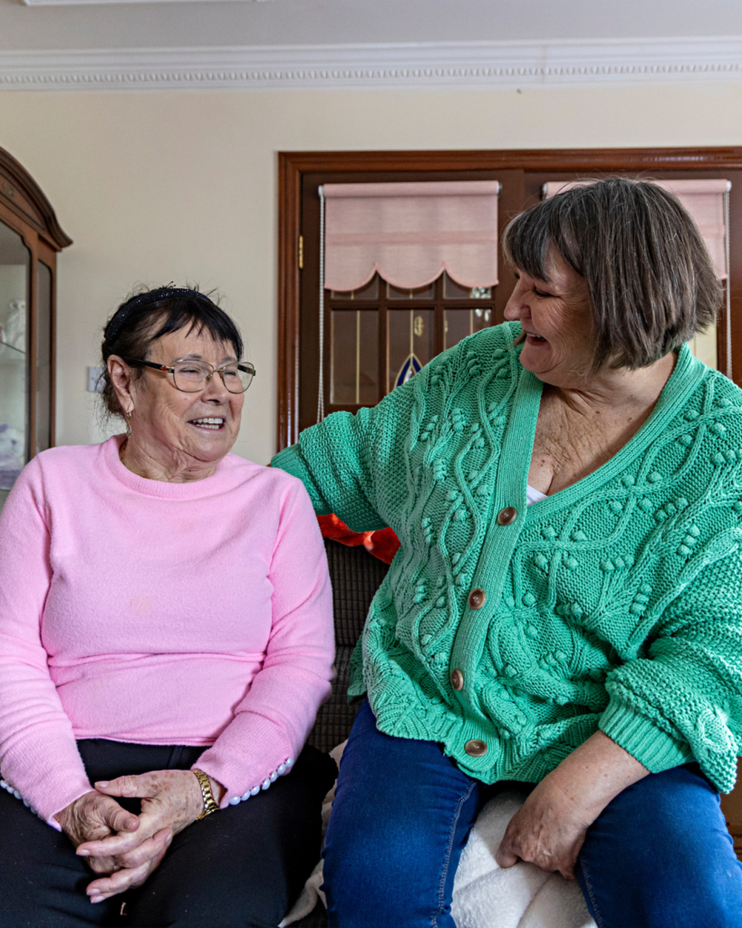 Two women laughing together on a sofa in a bright sitting room, one in pink, the other in a green cardigan