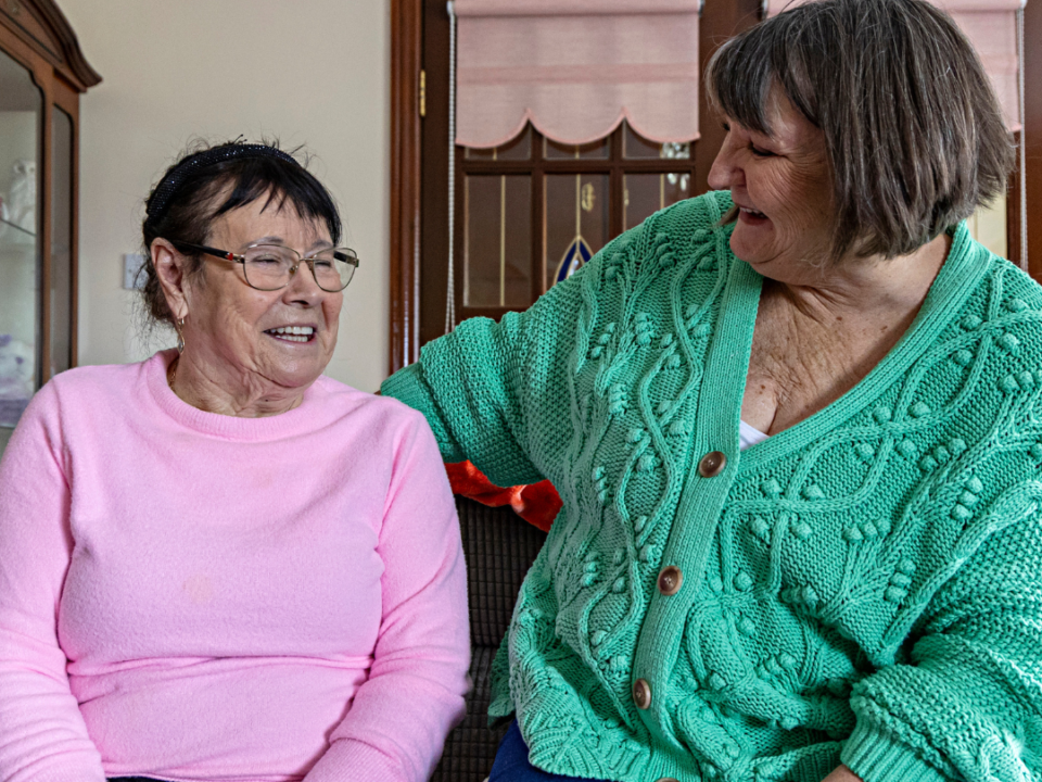 Two women laughing together on a sofa in a bright sitting room, one in pink, the other in a green cardigan