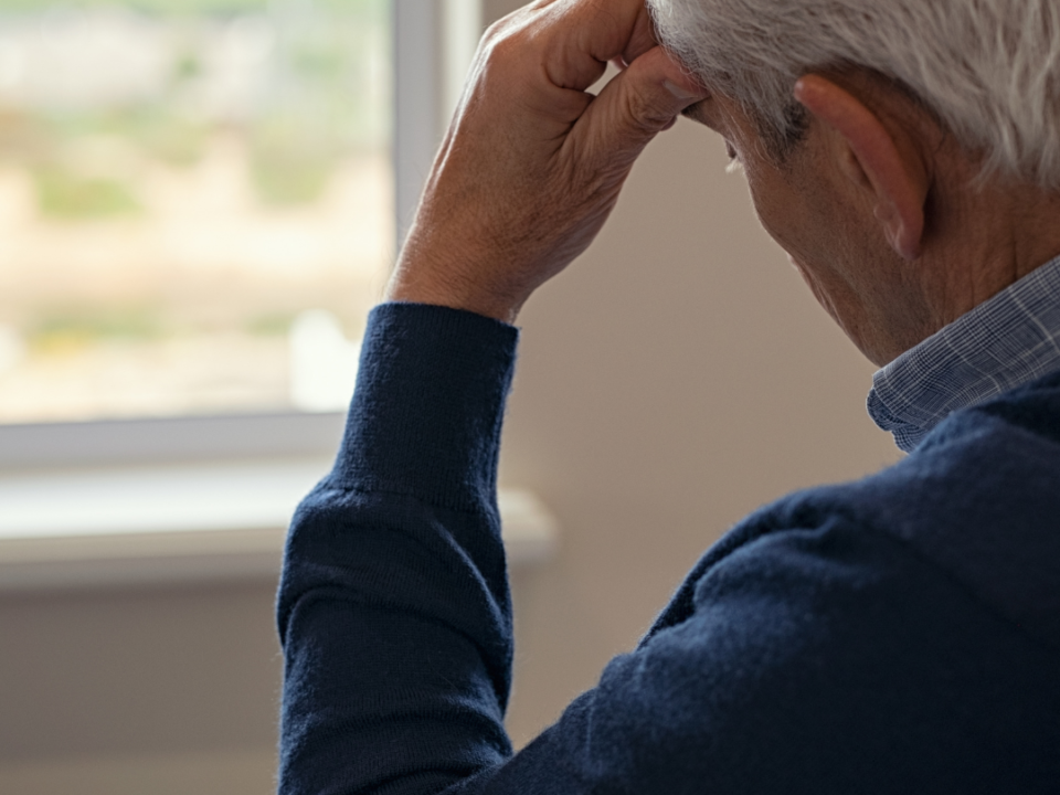 Older man in a navy jumper sits by a window with his hand to his forehead, looking downcast