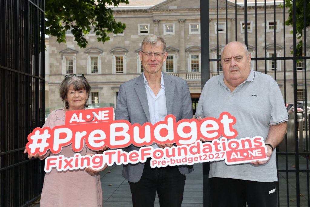 Three people outside Leinster House holding an ALONE sign reading '#PreBudget FixingTheFoundations Pre-budget2027'