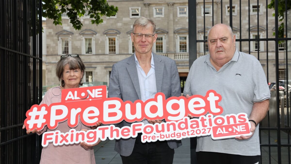 Three people outside Leinster House holding an ALONE sign reading '#PreBudget FixingTheFoundations Pre-budget2027'