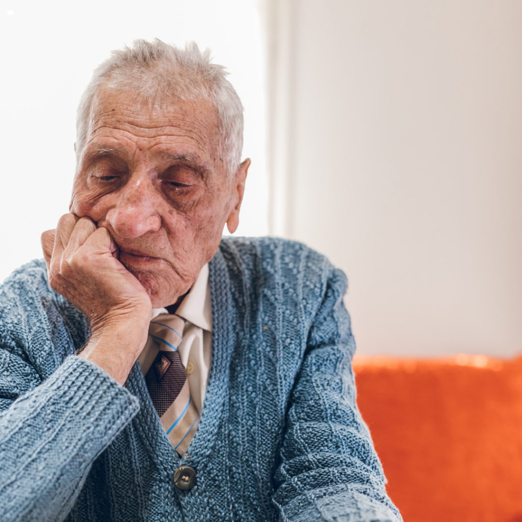 Older man in a blue knitted cardigan and tie sitting alone, resting his chin on his hand and looking down