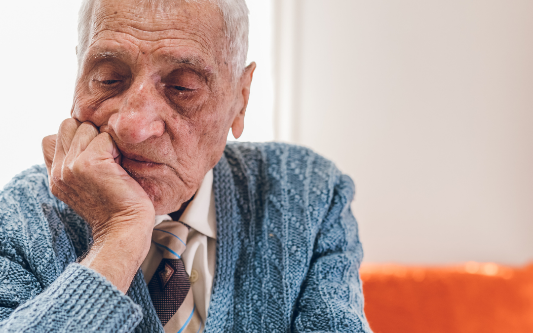 Older man in a blue knitted cardigan and tie sitting alone, resting his chin on his hand and looking down