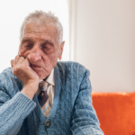 Older man in a blue knitted cardigan and tie sitting alone, resting his chin on his hand and looking down