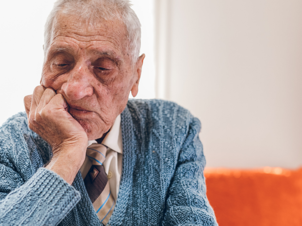 Older man in a blue knitted cardigan and tie sitting alone, resting his chin on his hand and looking down