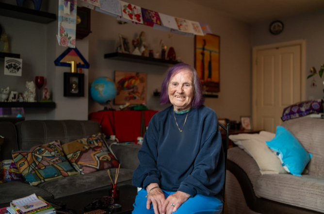 Older woman with purple-dyed hair smiling in her sitting room, surrounded by artwork and cushions