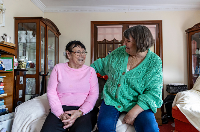 An older woman in pink and a woman in a green cardigan sitting together, laughing, in a sitting room at home